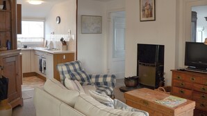A living room with a sofa, a television, a wooden cabinet, and a kitchen area with a stove and sink. - 4 bedroom accommodation in Pagham, near Chichester (Pagham, near Chichester)
