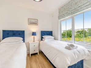 A double room with two beds, a nightstand with a lamp, a framed picture on the wall, and a window with a view of greenery.