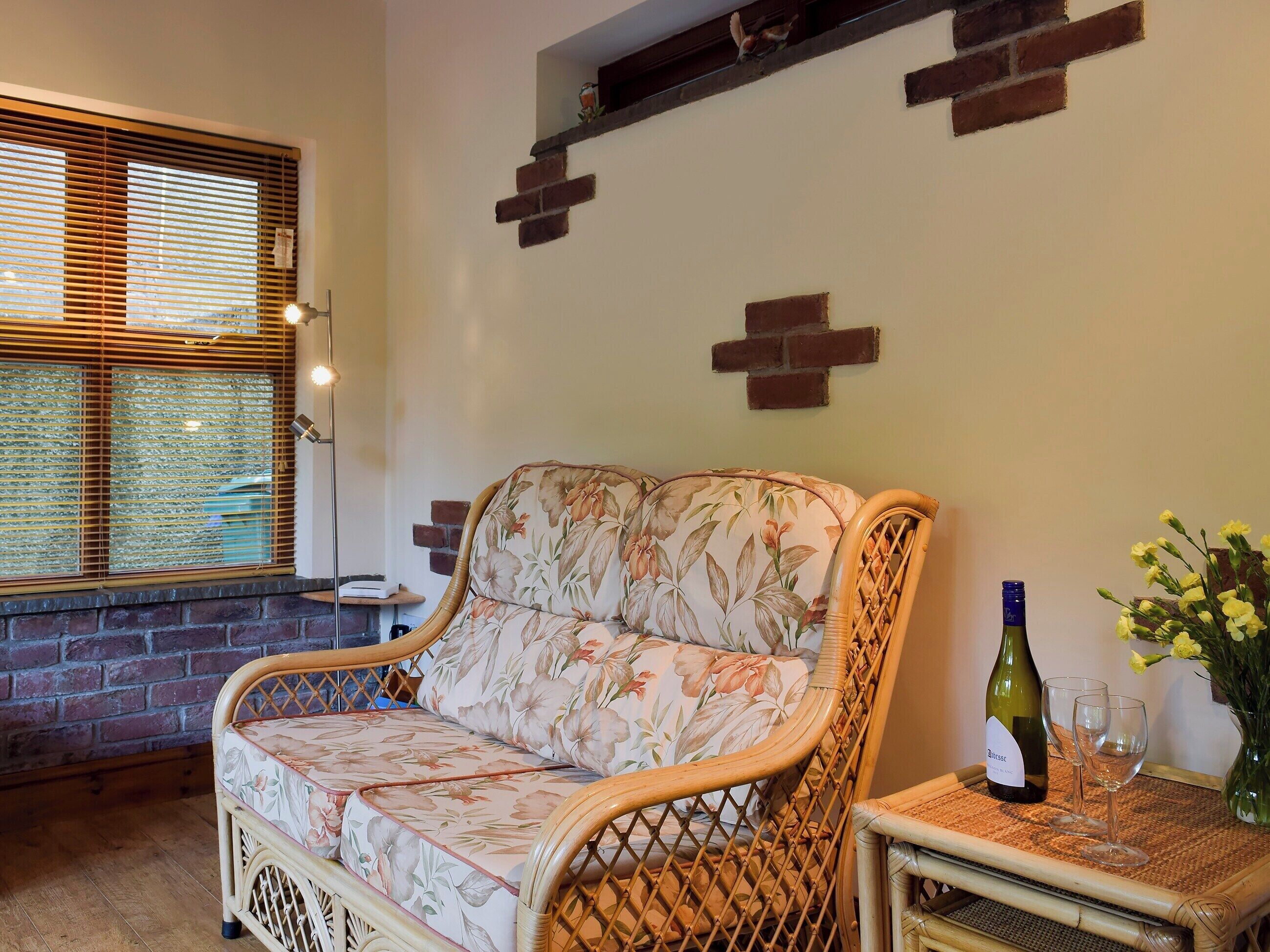A wicker sofa with floral upholstery, a small table with a wine bottle and glasses, and a brick wall with a shelf.