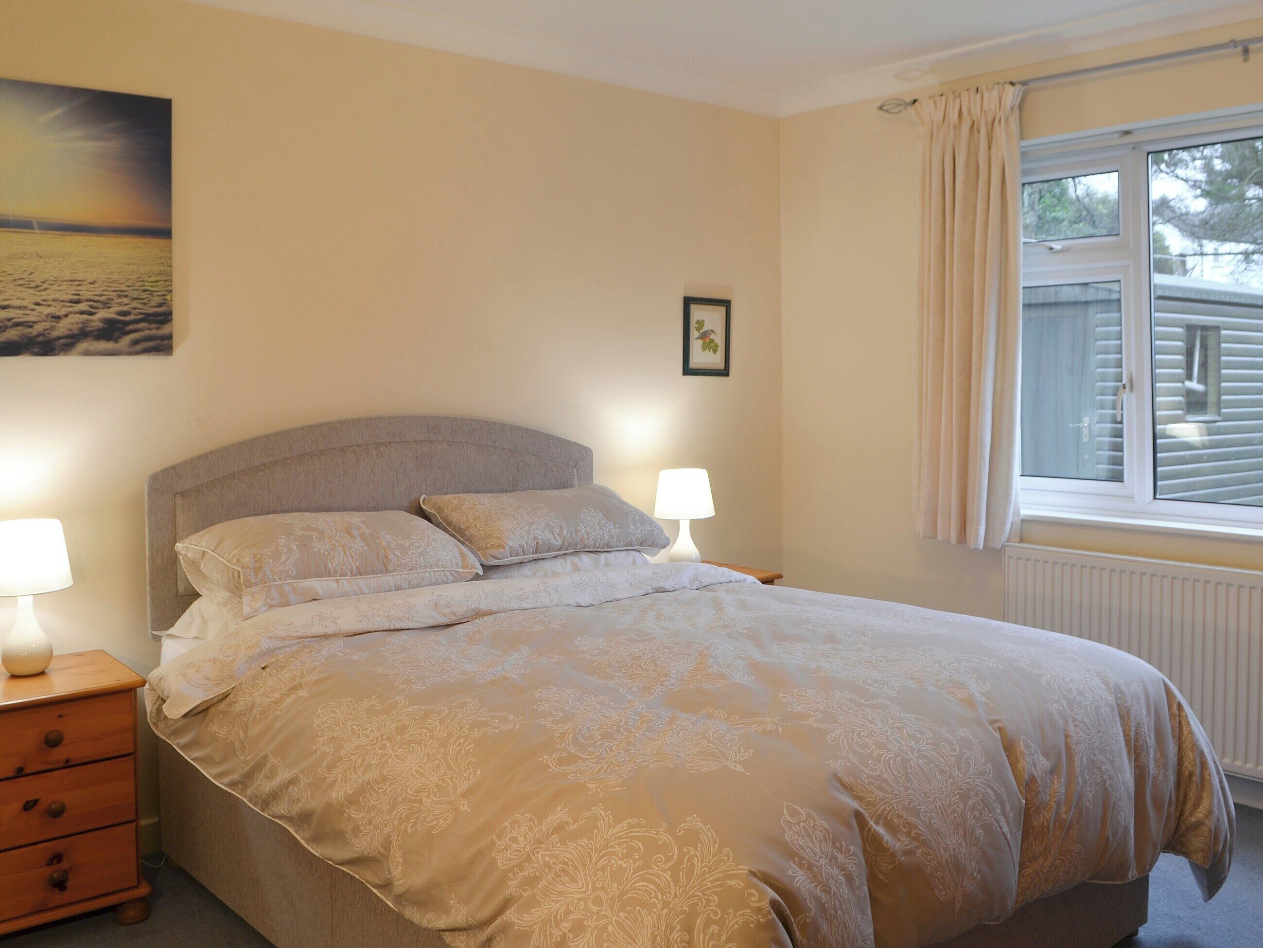 A bedroom with a bed, two bedside lamps, a nightstand, a framed picture on the wall, and a window with curtains.