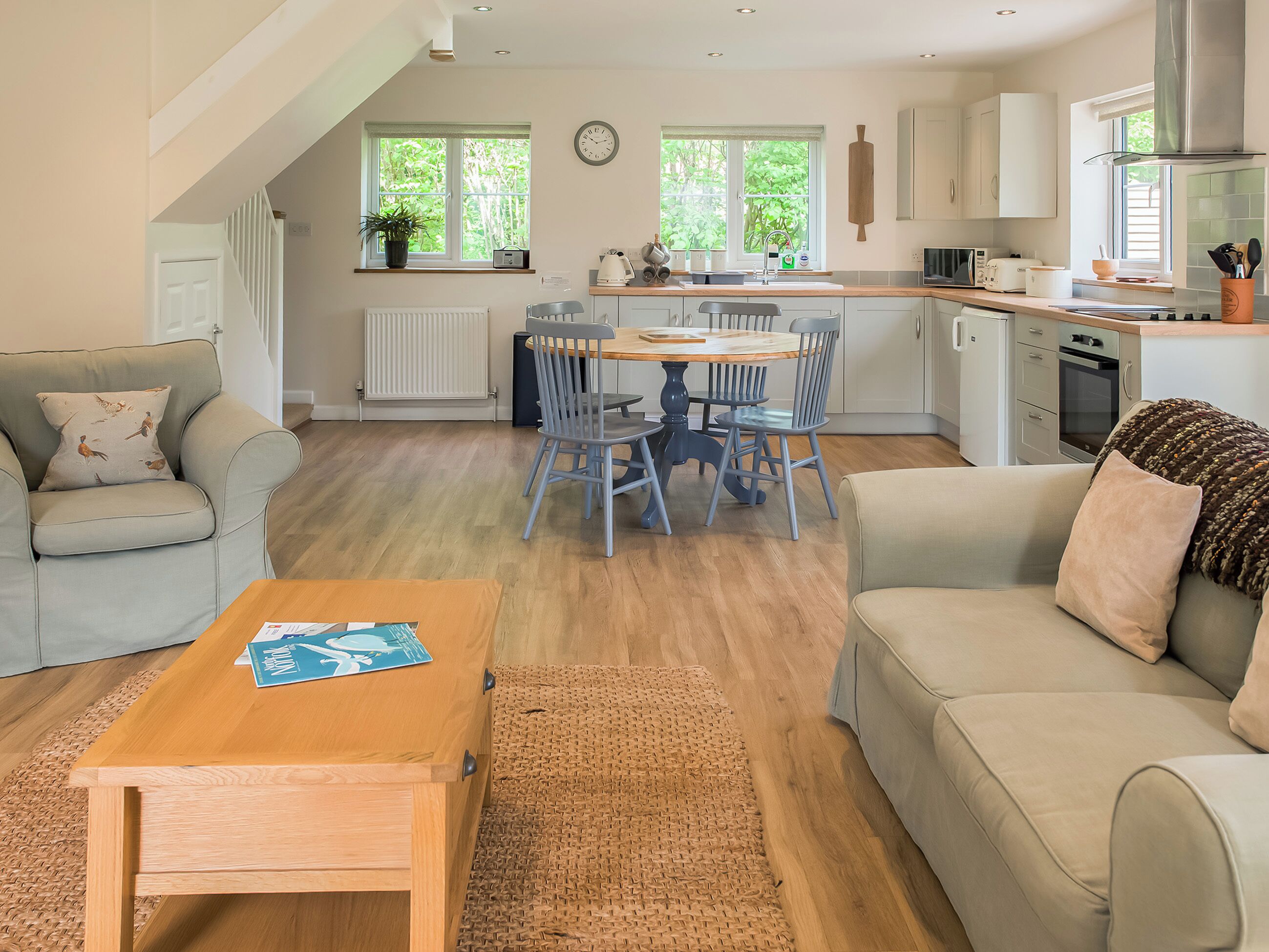 A modern kitchen with a dining area, wooden flooring, and a view of greenery outside.