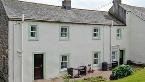 A two-story house with a stone roof, multiple windows with green frames, and a small outdoor seating area with a table and chairs. - 2 bedroom accommodation in Caldbeck, near Keswick (Caldbeck, near Keswick)