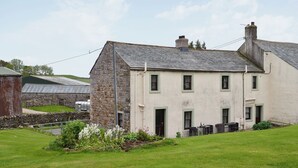 A two-story house with a stone facade and a sloped roof, surrounded by a well-maintained lawn and garden. - 2 bedroom accommodation in Caldbeck, near Keswick (Caldbeck, near Keswick)