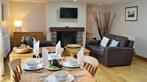 A dining area with a wooden table set for four, a fireplace, a leather sofa, and a television mounted on the wall. - 2 bedroom accommodation in Allonby, near Silloth (Allonby, near Silloth)