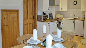 A compact kitchen with white cabinets, a wooden door, and a dining area set for four with white tableware. - 2 bedroom accommodation in Allonby, near Silloth (Allonby, near Silloth)