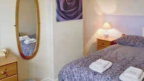 Un dormitorio con cama, mesita de noche con lámpara, cómoda, espejo y un cuadro de flores en la pared.
