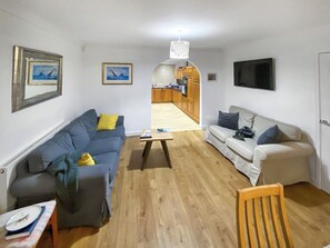 A living room with a sofa, a coffee table, and a TV. There is a kitchen visible in the background.