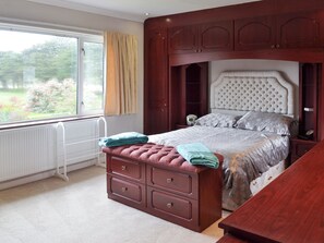 A bedroom with a large window, a bed with a headboard, a dresser with drawers, and a bedside table.