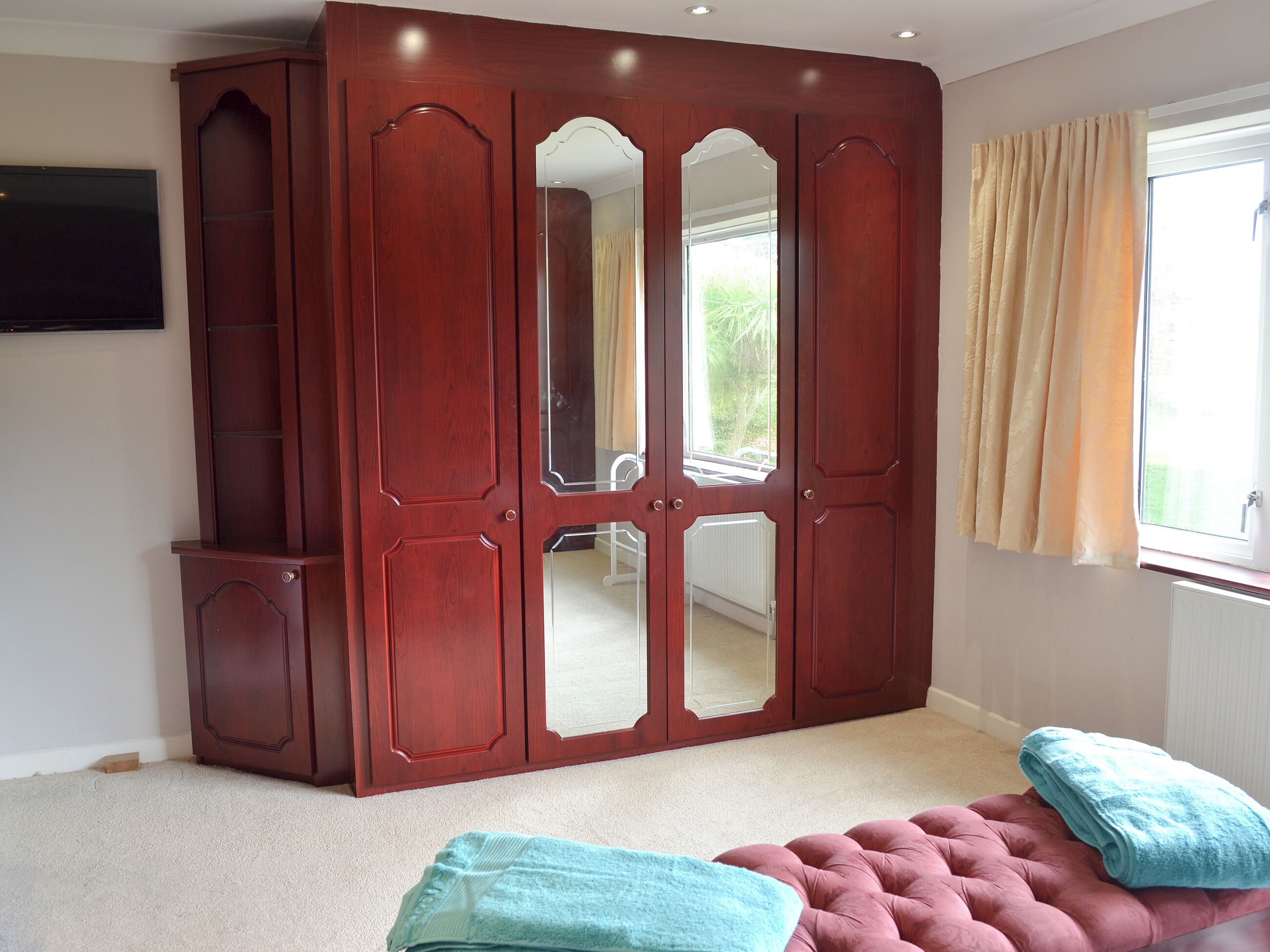 A bedroom with a large wooden wardrobe, a pink tufted ottoman, and a window with curtains.