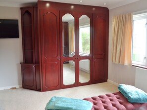 Ein Schlafzimmer mit großem Holzschrank, einem gepolsterten Ottoman in Rosa und einem Fenster mit Vorhängen.