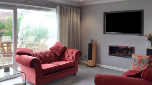 A living room with a red sofa, a television, a fireplace, and a large window. - 4 bedroom accommodation in Illogan, near Redruth (Illogan, near Redruth)