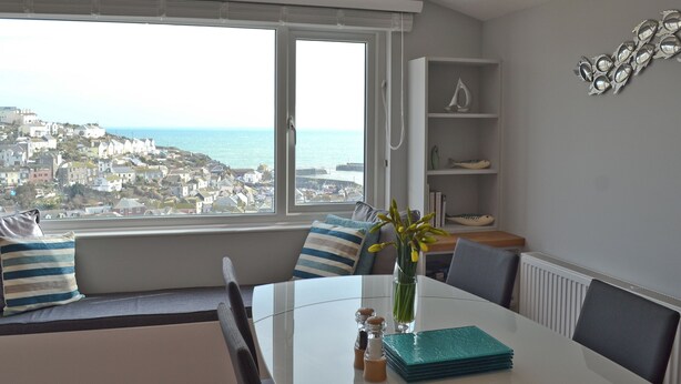 A dining area with a view of a coastal town, a table with a vase of flowers, and a shelf with decorative items. - 3 bedroom accommodation in Mevagissey (Mevagissey)