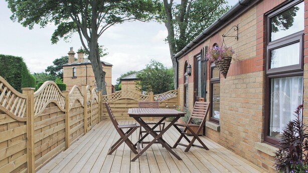 A wooden deck with a table and chairs, a brick building with a window, and a wooden fence. - 4 Eldin Hall Cottages (Scarborough)