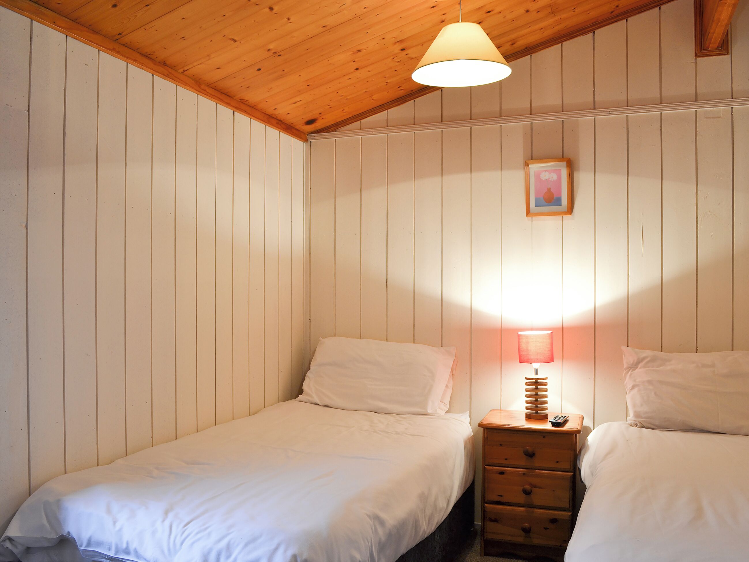 Een slaapkamer met twee eenpersoonsbedden, een houten hoofdbord, een nachtkastje met een lamp en een lijst met een afbeelding aan de muur.