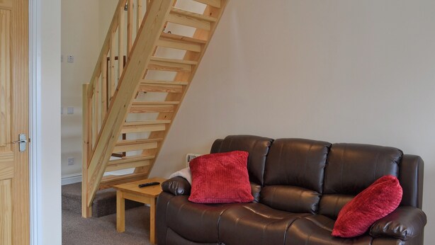A room with a brown leather sofa, two red cushions, a wooden staircase, and a small wooden table with a remote control. - 2 bedroom accommodation in Toynton-All-Saints, near Spilsby (Toynton-All-Saints, near Spilsby)