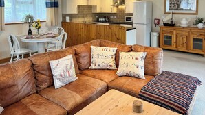 Ein gemütliches Wohnzimmer mit einem braunen Ledersofa, einem Holztisch und gemusterten Kissen. Im Hintergrund ist eine Essecke mit einem weißen Tisch und Stühlen zu sehen.