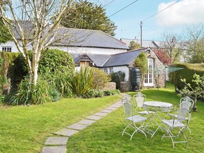 A garden with a stone pathway leading to a white metal table and chairs, surrounded by greenery and a stone building with a grey roof.