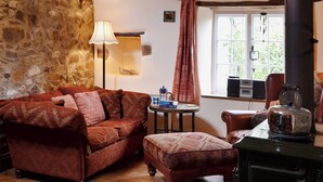 Una habitación acogedora con pared de piedra, suelo de madera, sofá estampado, una mesita redonda con libros y una tetera, y una ventana con cortinas.