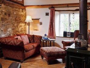 A cozy room with a stone wall, a wooden floor, a patterned sofa, a small round table with books and a kettle, and a window with curtains.
