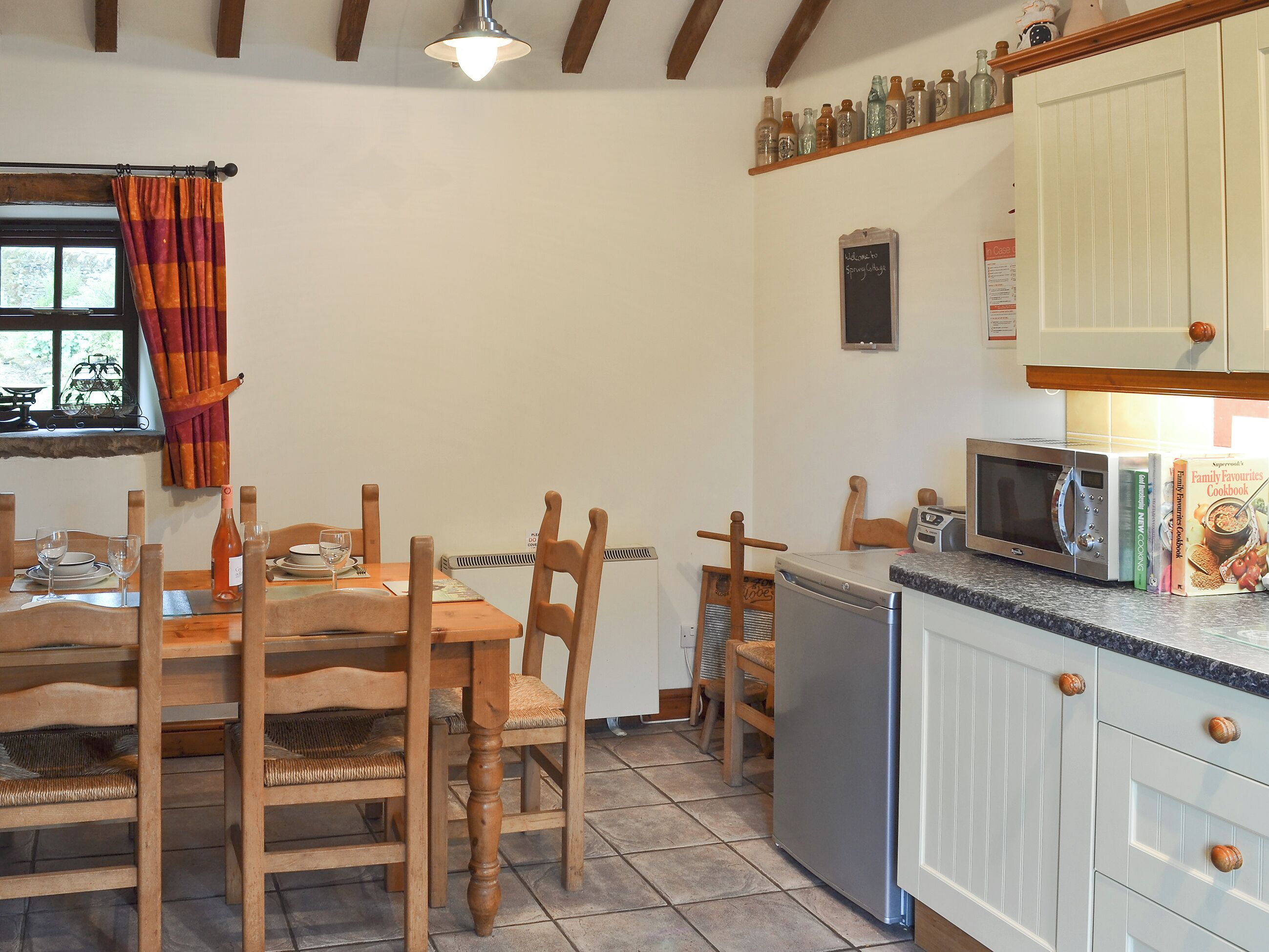 A kitchen with wooden chairs, a dining table, a microwave, and a shelf with bottles.