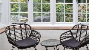 A room with two wicker chairs and a small glass table, large windows with white frames, and a view of greenery and a building outside. - 3 bedroom accommodation in Harmans Cross, near Swanage (Harmans Cross, near Swanage)