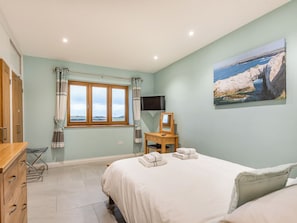A bedroom with a bed, a desk with a chair, a television, and a painting of a coastal scene.
