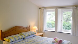 A bedroom with a wooden bed, a bedside table with a lamp, and two windows with curtains. - 2 bedroom accommodation in St Kew, near Port Isaac (St Kew, near Port Isaac)