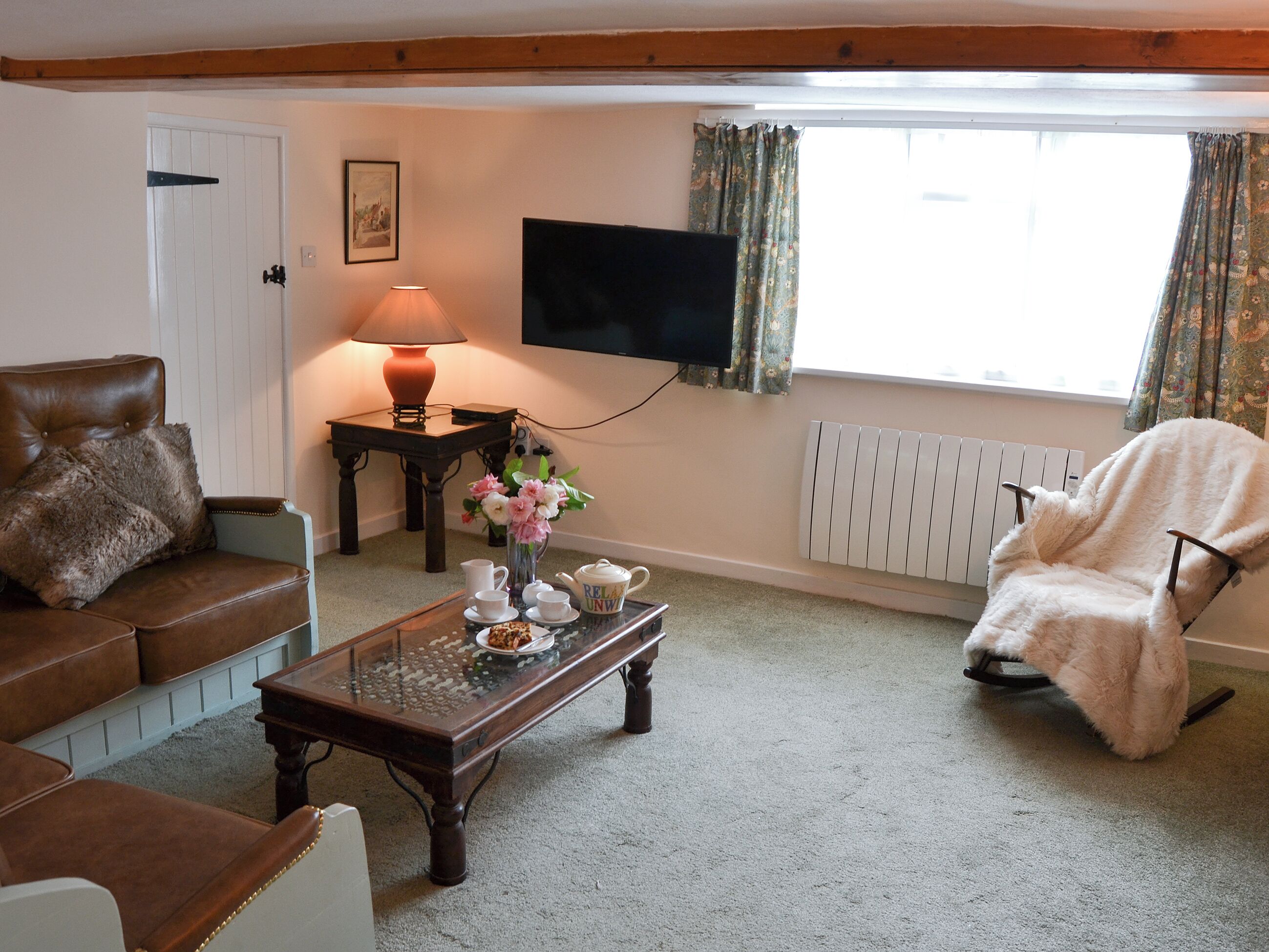 A living room with a brown leather sofa, a glass coffee table with tea set, a white armchair, a lamp, a TV, and a radiator.