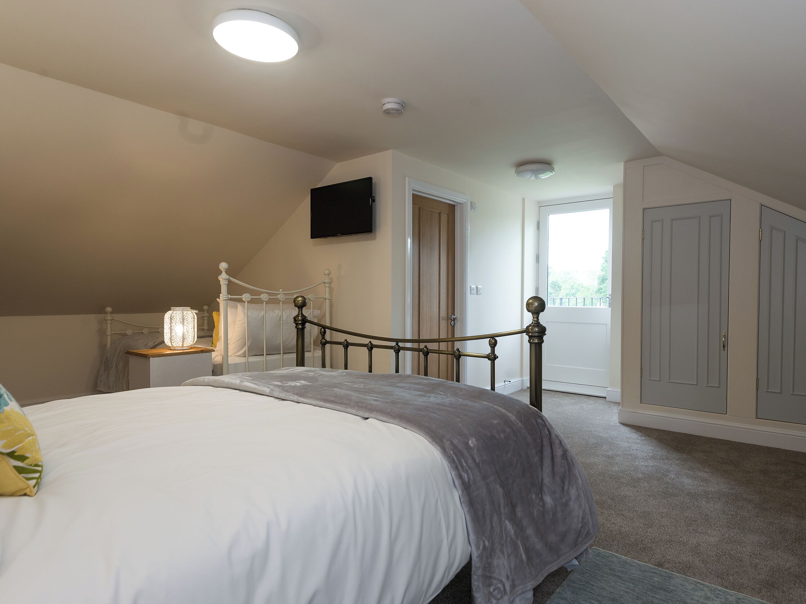 A bedroom with a bed, a bedside table, a lamp, a television mounted on the wall, and a door leading to another room.