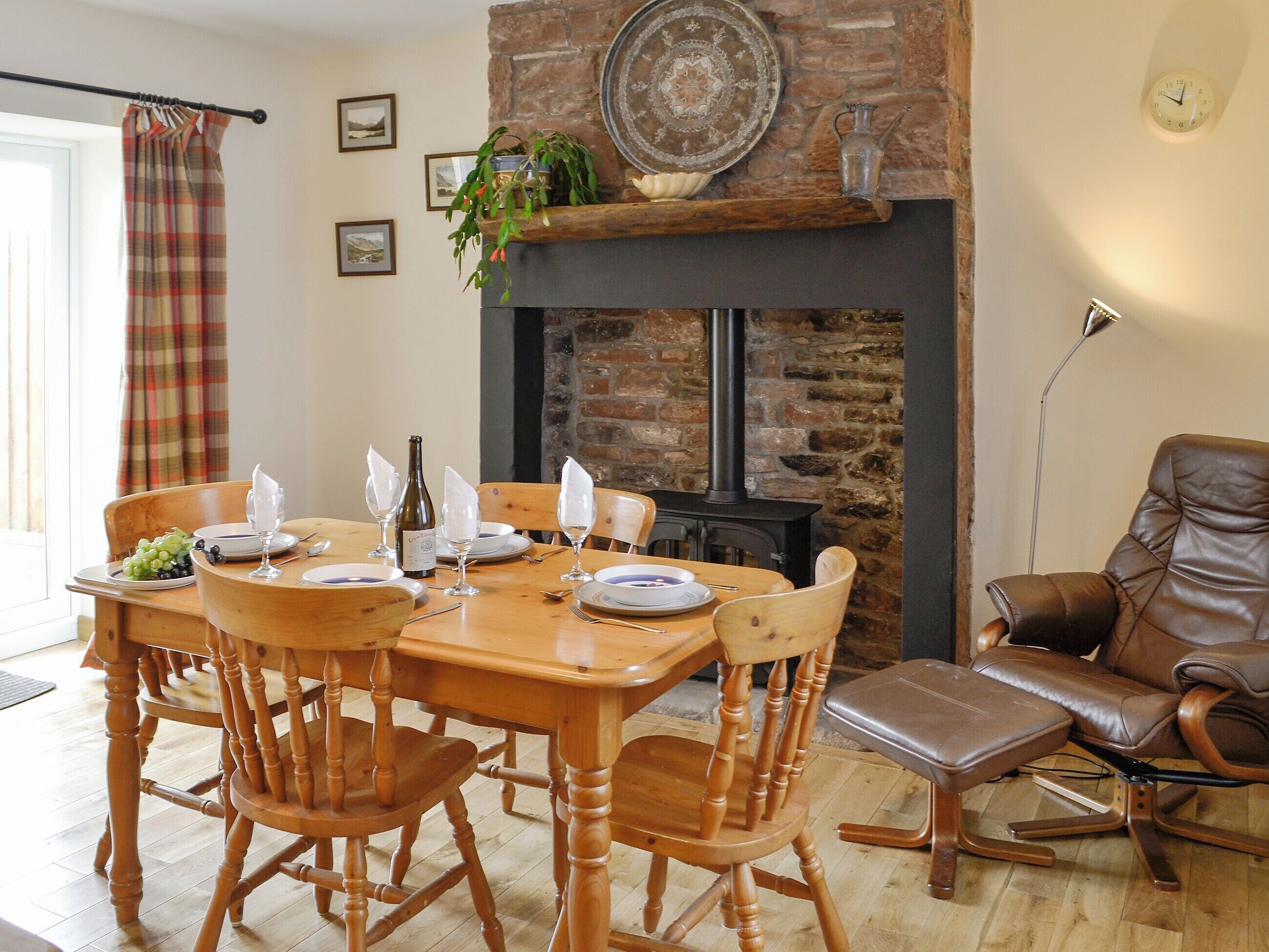 A dining area with a wooden table set for four, surrounded by wooden chairs. A fireplace with a stone facade is visible in the background.