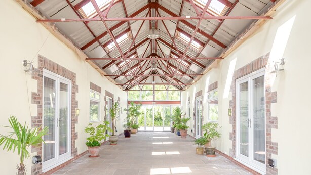 Un couloir couvert, doté d’un sol carrelé, de plantes en pot et de portes vitrées de part et d’autre.