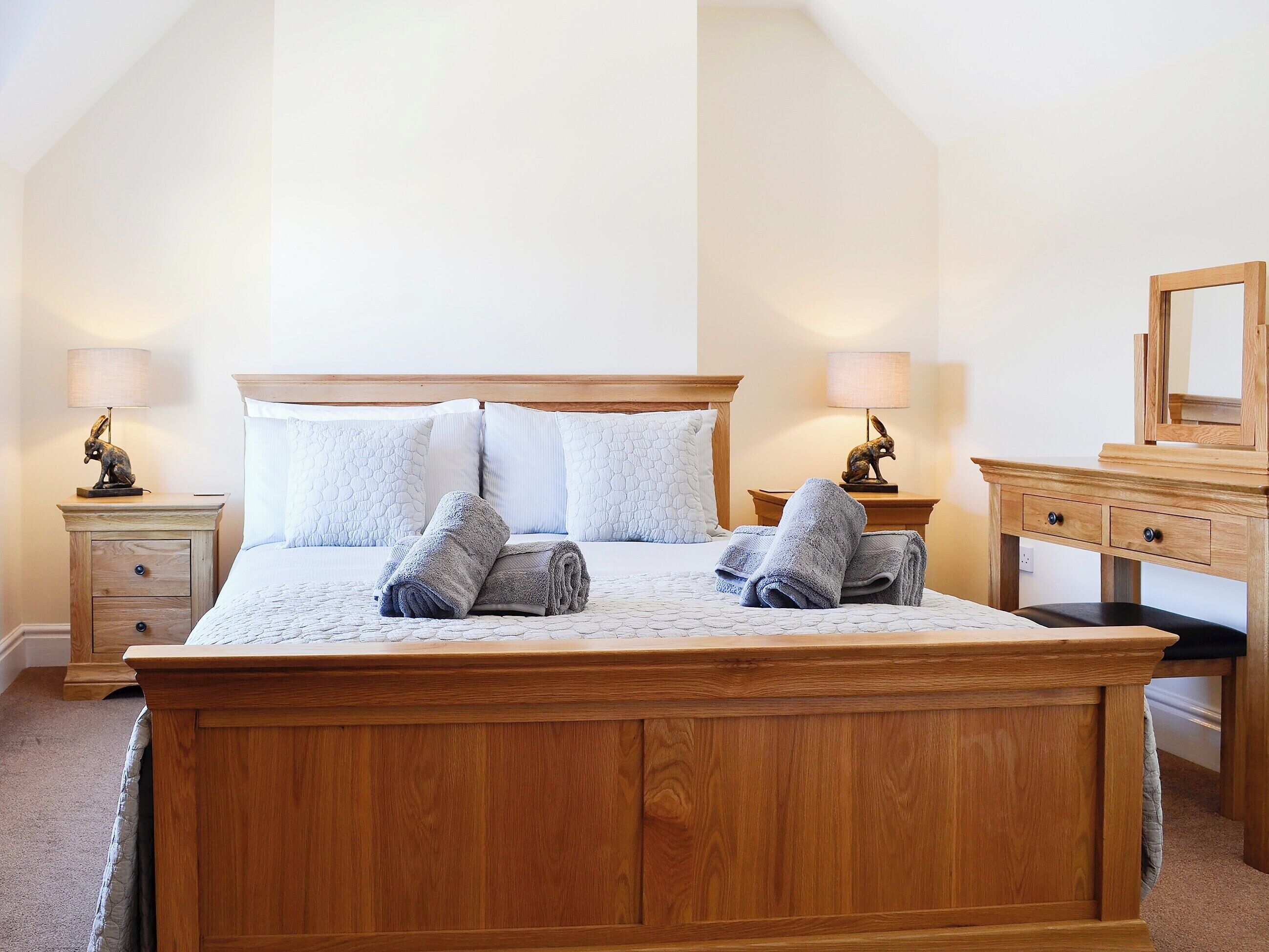 A neatly made bed with pillows and rolled towels, a wooden nightstand with a lamp, and a dresser with a mirror.