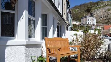 A wooden bench outside a white building with multiple windows, a stone wall, and a view of a hillside with buildings in the background.