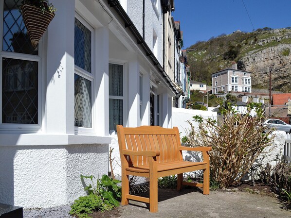 A wooden bench outside a white building with multiple windows, a stone wall, and a view of a hillside with buildings in the background.