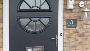 Una puerta gris oscuro con un panel de vidrio, un timbre y una placa con el número de la casa.