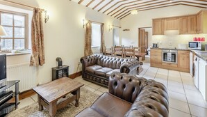 A cozy living room with a leather sofa, wooden dining table, and a fireplace. The kitchen area is visible with wooden cabinets and modern appliances.