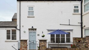 Une maison blanche à deux étages avec une porte bleue, un petit balcon agrémenté d’un parasol bleu et un mur en briques.