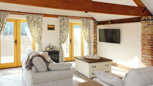 A cozy living room with a sofa, a coffee table, and a television mounted on a brick wall. There is a wooden ceiling with exposed beams, and a bicycle is visible near the door. - 3 bedroom accommodation in Tickton, near Beverley (Tickton, near Beverley)