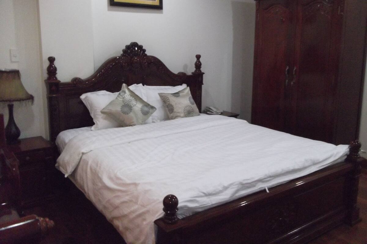 Quarto com cama de madeira, roupa de cama branca e mesa de cabeceira com abajur.