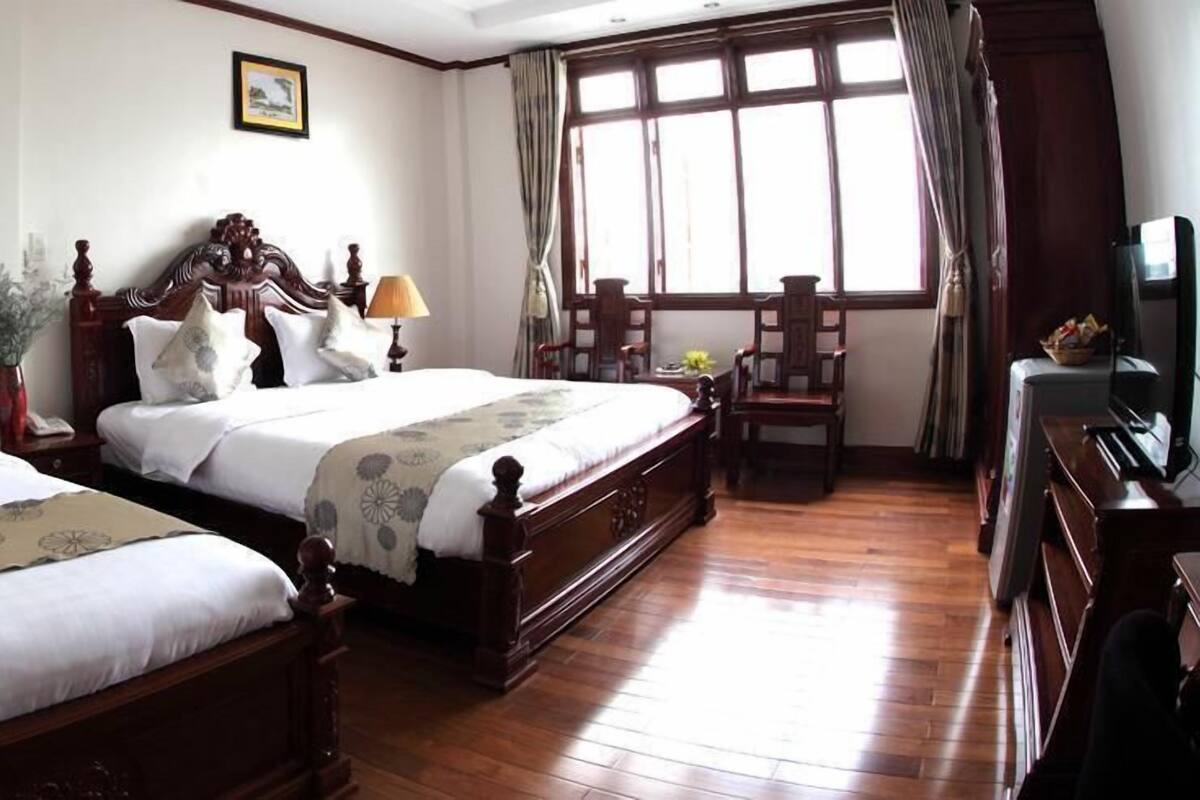 Quarto de hotel com duas camas de madeira, uma janela grande com cortinas, uma escrivaninha com abajur e um piano.