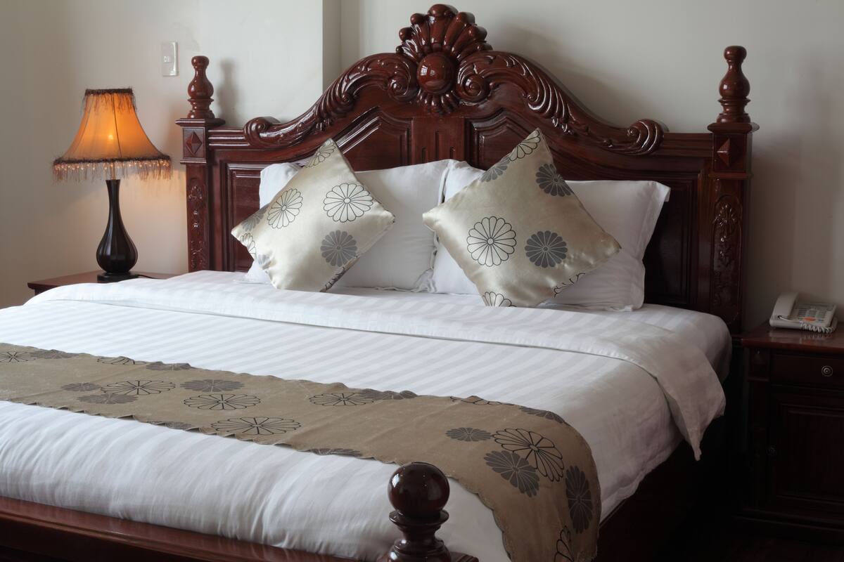 Cama bem arrumada com colcha de padrão floral, duas almofadas decorativas, abajur e mesa de cabeceira de madeira com telefone.