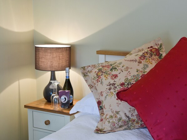 A bed with a floral pillow and a red blanket, a bedside table with a lamp, a bottle, and a glass.