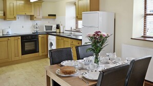 A modern kitchen with wooden cabinets, a built-in oven, microwave, and washing machine. There is a dining table set with plates, cups, and a vase of flowers. - 2 bedroom accommodation in Northlew, near Tavistock (Northlew, near Tavistock)