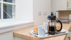 Una cocina con ventana, una cafetera, un croissant y dos tazas blancas sobre una bandeja.