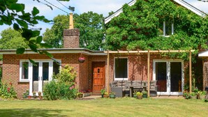 Une maison en briques avec une cheminée, une véranda avec des sièges d’extérieur et un jardin agrémenté de plantes en pot.