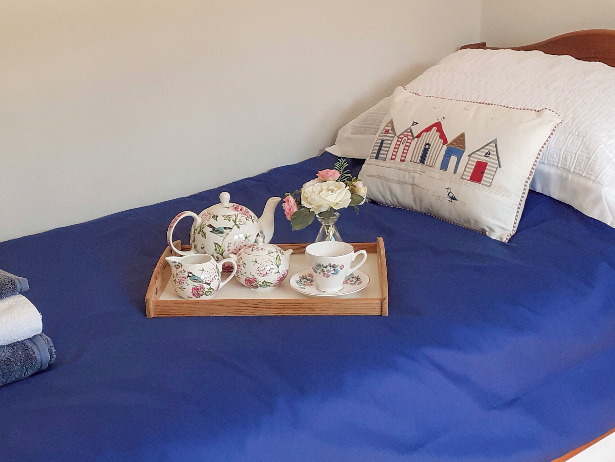 Ein Bett mit blauer Bettdecke, ein Holztablett mit Teekanne mit Blumenmuster, eine Tasse und eine Vase mit Blumen.