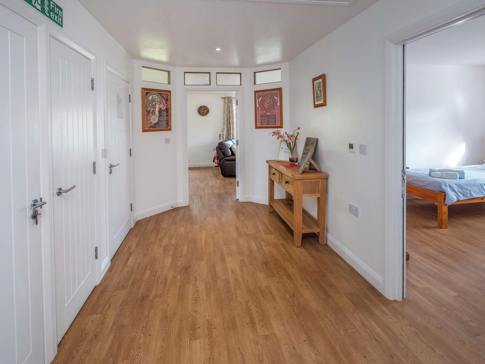 Un couloir avec des portes blanches, un plancher en bois, un bureau avec un vase, et un lit à l’arrière-plan.