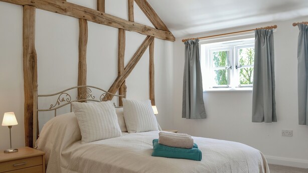 A bedroom with a wooden ceiling, a bed with white bedding, a nightstand, a lamp, and a window with curtains. - 4 bedroom accommodation in Ashperton, near Ledbury (Ashperton, near Ledbury)