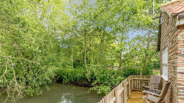 A wooden deck with chairs overlooking a river, surrounded by lush greenery. - COTTAGE IN THE POND W43212 (Garton, near Hornsea)
