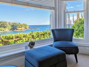 A room with a large window offering a view of a coastal landscape, a blue chair, and a small ottoman. - Seascape - St Catherine's Court (Fowey)